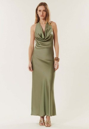 Femme debout portant une robe sans manches en satin vert olive avec un col bénitier, des sandales dorées et un bracelet doré épais au poignet droit.