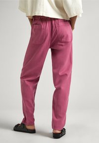 Persona vistiendo pantalones rosas de corte relajado con bolsillos parche, zapatos negros sin cordones con lazos y una blusa cremosa texturizada, de pie frente a un fondo claro.