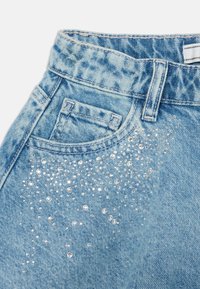 Los jeans de mezclilla azul claro presentan una textura desgastada y cuentas decorativas de pedrería transparente dispuestas en un degradado en el área del bolsillo delantero.