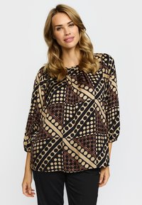 Kvinna med vågigt blont hår som bär en svart, beige och brun blus med geometriskt mönster, trekvartsärmar och svarta byxor.