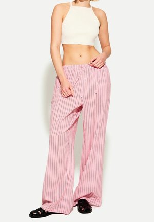 Kobieta w białym, krótko skrojonym topie i luźnych różowych spodniach w paski, poprawiająca pasek, stojąca na białym tle.