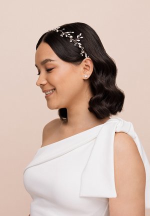 Abito bianco off-shoulder con un design drappeggiato morbido e un dettaglio annodato sulla spalla. Una fascia per capelli in argento e perle aggiunge accenti decorativi.