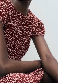 Robe rouge à pois blancs, manches courtes bouffantes, corsage froncé. Gros plan sur un bras reposant sur la robe, texture de peau lisse visible.