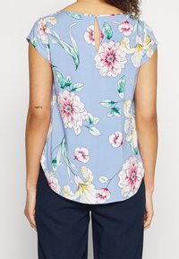 Blusa floreale in tessuto azzurro chiaro, con fiori rosa e gialli, maniche corte e un dettaglio a chiave nella parte posteriore. Orlo arrotondato.