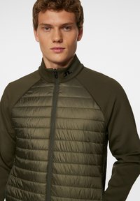 Miesmalli, jolla on lyhyet, kiharat hiukset, pukeutuneena oliivinvihreään vetoketjulliseen takkiin, jossa on kirjailtu etuosa ja sileät hihat, poseeraa vaaleaa taustaa vasten.