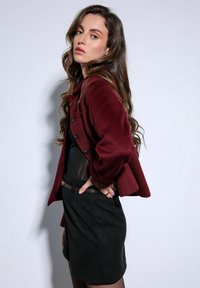 Bordeauxwollen jas met een cropped design, grote knopen en wijde mouwen, gedragen over een zwart nauwsluitend topje en rok.