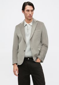 Světle šedý kordový blazer s klopami, jedním knoflíkem a bočními kapsami, v kombinaci se svetrem se zipem v tónu světle šedé a tmavými kalhotami.