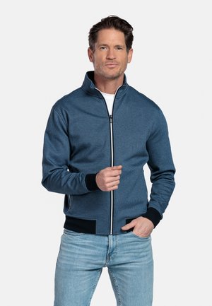 Man draagt een blauwe jas met ritssluiting, zwarte manchetten en kraag, wit shirt eronder en lichtblauwe spijkerbroek, staand tegen een effen achtergrond.