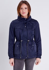 Giacca leggera blu navy con colletto alto, vita con cordino, due tasche sul petto e accenti metallici. Texture del tessuto liscia.