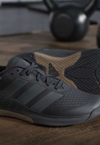 Μαύρα αθλητικά παπούτσια Adidas με πλέγμα και συνθετικό επάνω μέρος, τοποθετημένα σε ξύλινο πάτωμα με θολωμένα βαράκια στο φόντο.
