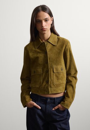 Junge Frau trägt eine olivgrüne Wildlederjacke mit Knopftaschen und dunkelblaue Jeans, steht mit den Händen in den Taschen.
