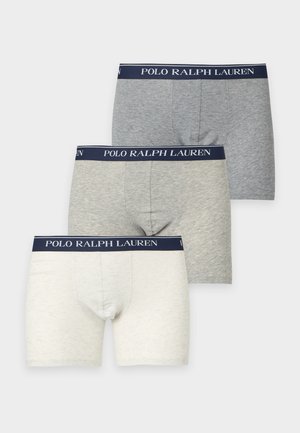 Wysoki, szczupły mężczyzna stojący boso, ubrany w białe bokserki z granatową gumką z napisem Polo Ralph Lauren na jednolitym białym tle.