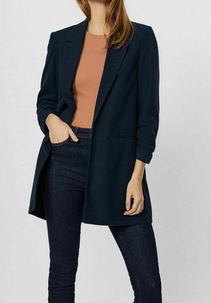 Woman wearing dark blue blazer over brown top and dark jeans, with one hand in pocket and the other relaxed by her side.