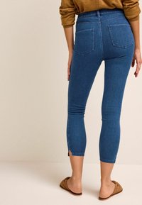 Jean capri en denim bleu taille haute avec deux poches arrière et des fentes latérales à l'ourlet, porté avec une sandale slide beige sur un fond clair.