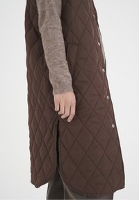 Brun quiltad väst med diamantmönster, sidofickor och tryckknappar. Buren över en ljusbrun tröja, som framhäver en lager-på-lager-textur.