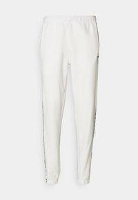 Bílé tepláky s elastickým pasem, zúženými nohavicemi a černými postranními pruhy s logo detaily. Hladký povrch, volný střih.