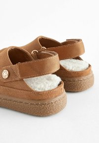 Sandales en suede marron avec des semelles intérieures en peluche blanche, des sangles en tissu ajustables et des semelles en caoutchouc texturé. Design simple avec des accents floraux.