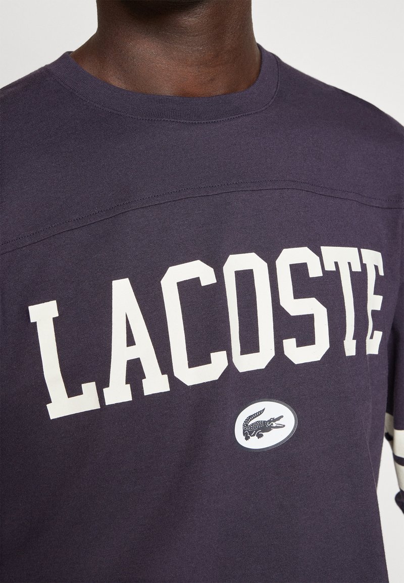 Close-up van een persoon in een donkerpaarse Lacoste-sweatshirt met grote witte "LACOSTE"-tekst en het krokodillenlogo van het merk eronder.