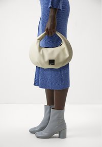 Krémová kabelka s zakřiveným tvarem a černou logovou nášivkou, spárovaná s modrými žebrovými šaty a šedými koženými kotníkovými botami na blokovém podpatku.