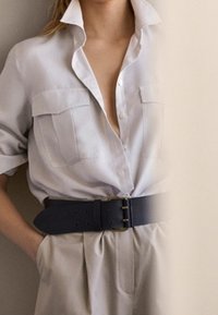 Jasnoszara koszula z kołnierzykiem z dwoma kieszeniami na piersi, połączona z beżowymi spodniami o wysokim stanie i szerokim czarnym paskiem. Prosty, dopasowany krój.