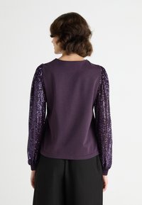 Violetinis džemperis su prigludusia nugara ir apimtais blizgučiais rankovėmis, turintis apvalią iškirptę ir rausvusiomis rankovėmis, sukurtas šiuolaikinei siluetei.