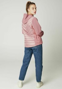 Lyserød polstret jakke med hætte, der har en ribbet tekstur på ærmerne og et chevron-design på ryggen, parret med blå jeans.