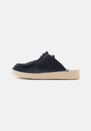 Navyblauwe suède instap-mules met mocassinnaaien, veterdetail bovenop en een beige rubberen zool, vanaf de zijkant weergegeven tegen een witte achtergrond.