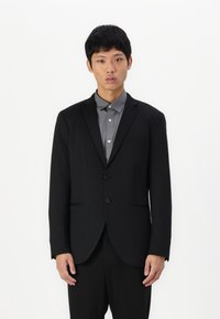 Čierny blazer so zapínaním na dva gombíky, výstužné klopy a predné vrecká, v kombinácii so šedou košeľou s golierom. Hladká textura, štruktúrovaný dizajn.