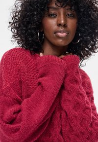 Czerwony dziergany sweter z fakturowanym wzorem, o luźnym kroju i oversize'owych rękawach, wykonany z miękkiego, przytulnego materiału.