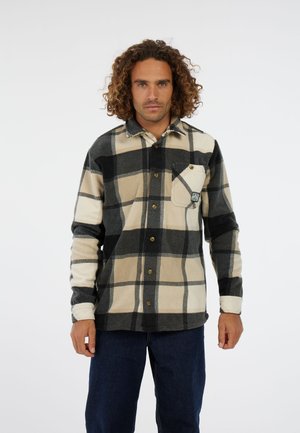 Man met krullend haar, gekleed in een beige, zwarte en crèmekleurige geruite overhemden met een borstzak, staande tegen een effen witte achtergrond.