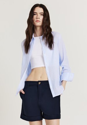 Camicia a righe blu chiaro e bianca con bottoni, top corto bianco e pantaloni corti blu navy con orli arrotolati. Il materiale sembra liscio; i dettagli dei bottoni sono visibili.