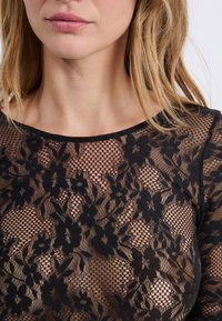 Top en dentelle noire avec des motifs floraux, présentant une texture transparente, un col rond et des manches longues, mettant en avant un design délicat et translucide.
