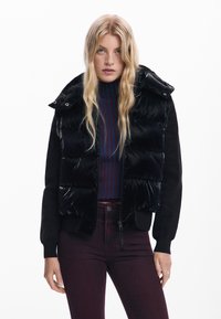 Piumino nero con finitura lucida, maniche nere in maglia, colletto alto, chiusura a zip, indossato sopra un maglione navy a righe e jeans bordeaux.