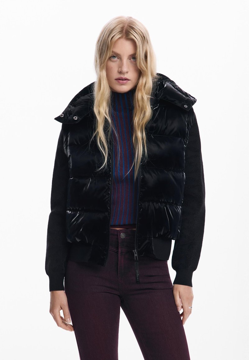 Piumino nero con finitura lucida, maniche nere in maglia, colletto alto, chiusura a zip, indossato sopra un maglione navy a righe e jeans bordeaux.