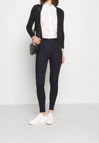 Czarny kardigan, biała koszula z abstrakcyjnym wzorem, ciemnogranatowe dopasowane spodnie oraz białe trampki. Prosty design z dopasowaną sylwetką.