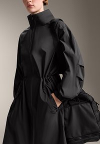 Zwarte jas met hoge kraag, elastische taille en oversized mouwen. Zijzakken en een gestructureerde zwarte tas met banden. Glad stof.