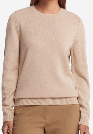 Femme portant un pull tricoté à manches longues beige et un pantalon marron, montrée de l'épaule aux hanches sur un fond blanc.