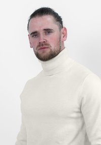 Beige coltrui gemaakt van zacht gebreid materiaal, met een hoge kraag en een aansluitend ontwerp, gedragen door een persoon voor een effen achtergrond.