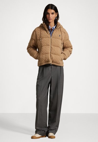 Tan gewatteerde jas met kabelbreipatroon, ritssluiting en capuchon. Gecombineerd met grijze broeken en beige sneakers, alles tegen een neutrale achtergrond.