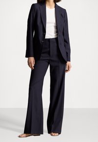 Kvinna klädd i en marinblå blazer och matchande vida byxor med en vit topp och öppna sandaler, stående mot en enklare bakgrund.