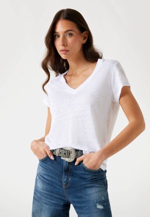 White v-neck t-shirt in lightweight fabric, paired with blue denim jeans and a black belt featuring silver embellishments and a decorative buckle.