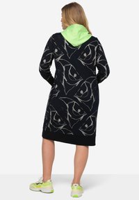 Robe hoodie noire à motifs avec capuche verte, ornée de motifs abstraits gris, à manches longues et à coupe décontractée, associée à des baskets blanches.