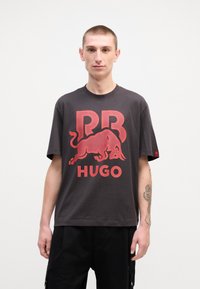 Dark gray t-shirt with a large red "RB" and bull graphic, and "HUGO" text in red. Unadorned short sleeves, made from soft cotton fabric.