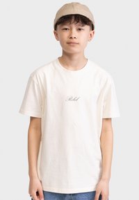 T-shirt en coton couleur crème avec un col rond, présentant un petit texte "Rebel" imprimé en lettres foncées au centre.