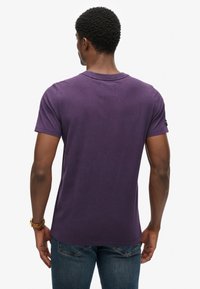 Fioletowa krótko rękawowa koszulka wykonana z bawełny, z okrągłym dekoltem i lekko wydłużonym tyłem. Brak widocznych wzorów lub akcentów.