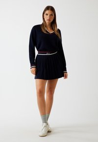 Marineblauwe trui met een V-hals en gestreepte accenten, gecombineerd met een marineblauwe plisséhart dat een gemarkeerde tailleband heeft. Draagt witte sneakers en grijze sokken.