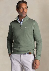 Groene kwart-rits pullover gemaakt van zachte stof, met een witte rits, naaddetails en gedragen met lichtgekleurde broeken en een gestreepte riem.