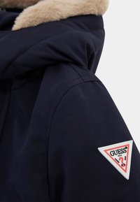 Jūrininkų striukė su dirbtinės kailių apykakle ir peties įvoku, kuriame pavaizduota raudona trikampio logotipas su tekstu "GUESS?" ir klausimo ženklu. Lygus audinys.