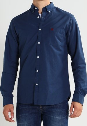 Homme portant une chemise bleu marine à boutons avec poche poitrine et un jean bleu foncé, debout devant un fond uni.