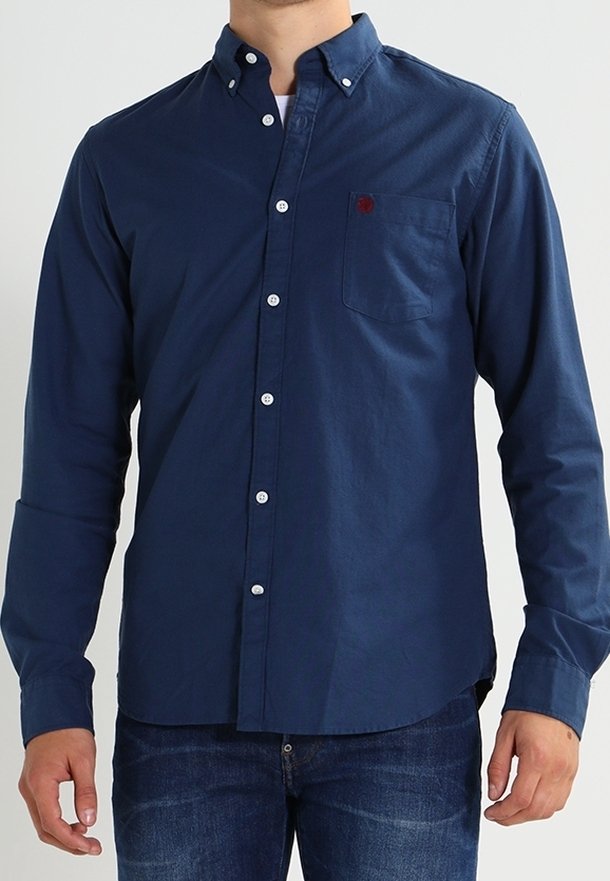Homme portant une chemise bleu marine à boutons avec poche poitrine et un jean bleu foncé, debout devant un fond uni.